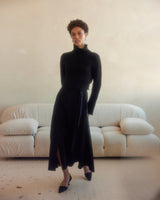 Model wearing a black ribbed cropped sweater with a black skirt, standing in a bright room.
