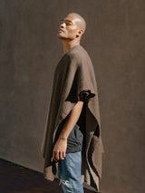 Model wearing a brown alpaca duster poncho, standing against a textured wall