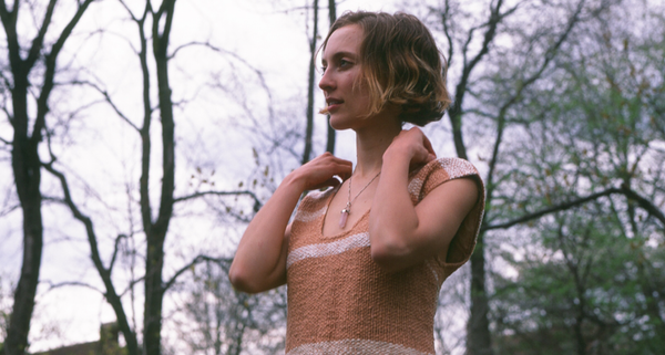 A woman in a knitted top standing outdoors with trees in the background, looking thoughtful.
