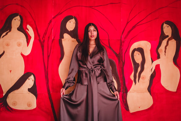 A woman in a gray dress poses in front of a red background with painted figures of women.