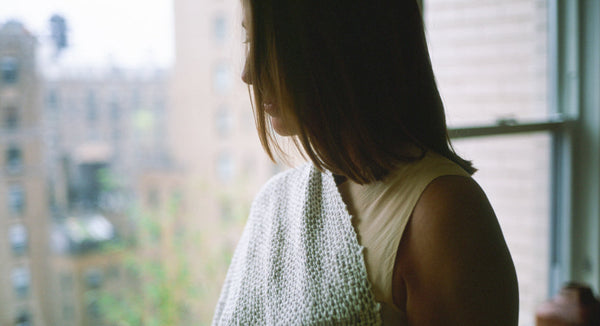 A woman with shoulder-length hair wearing a light top and holding a knitted shawl, standing by a window