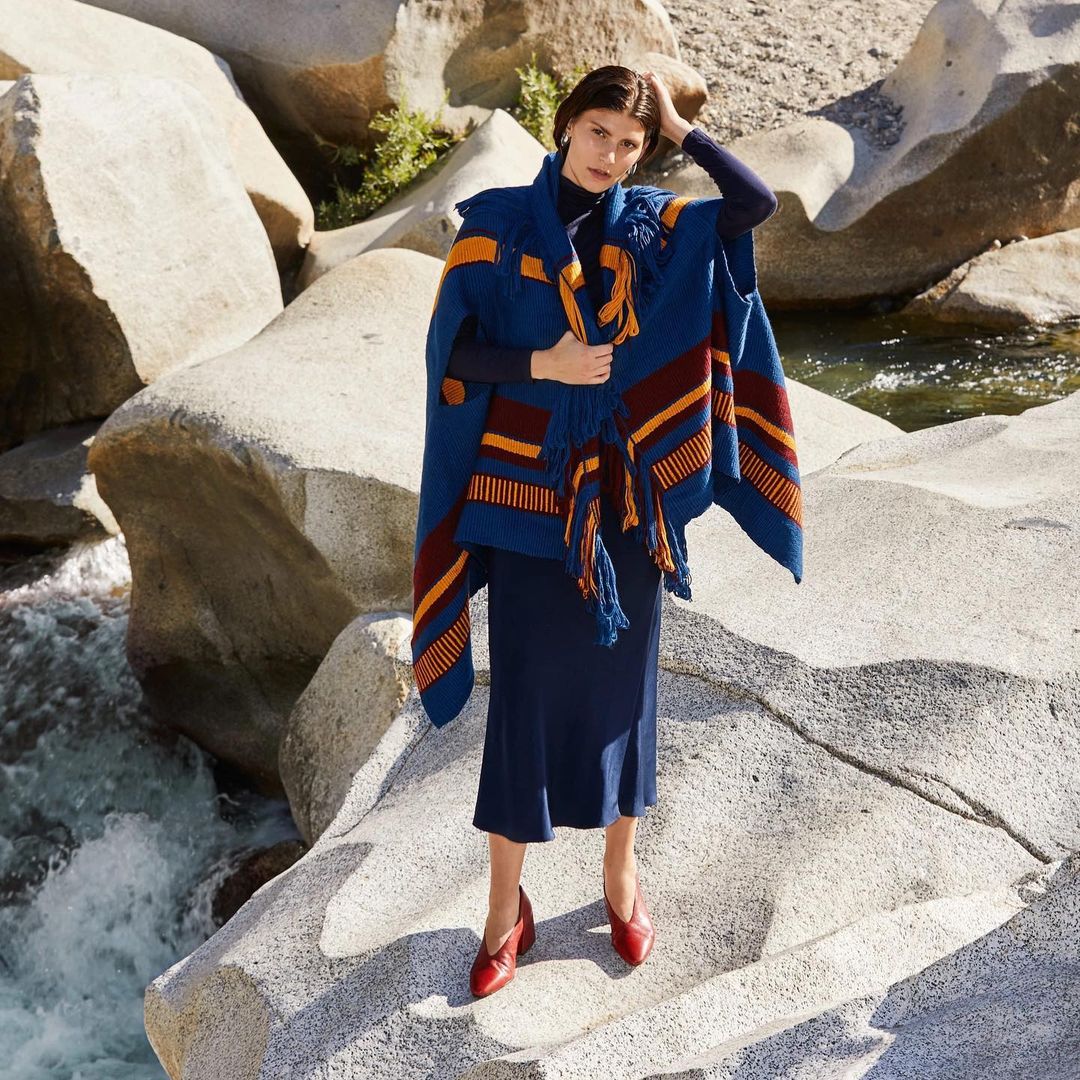 Model wearing a blue poncho with red and orange accents, standing on rocks by a stream.