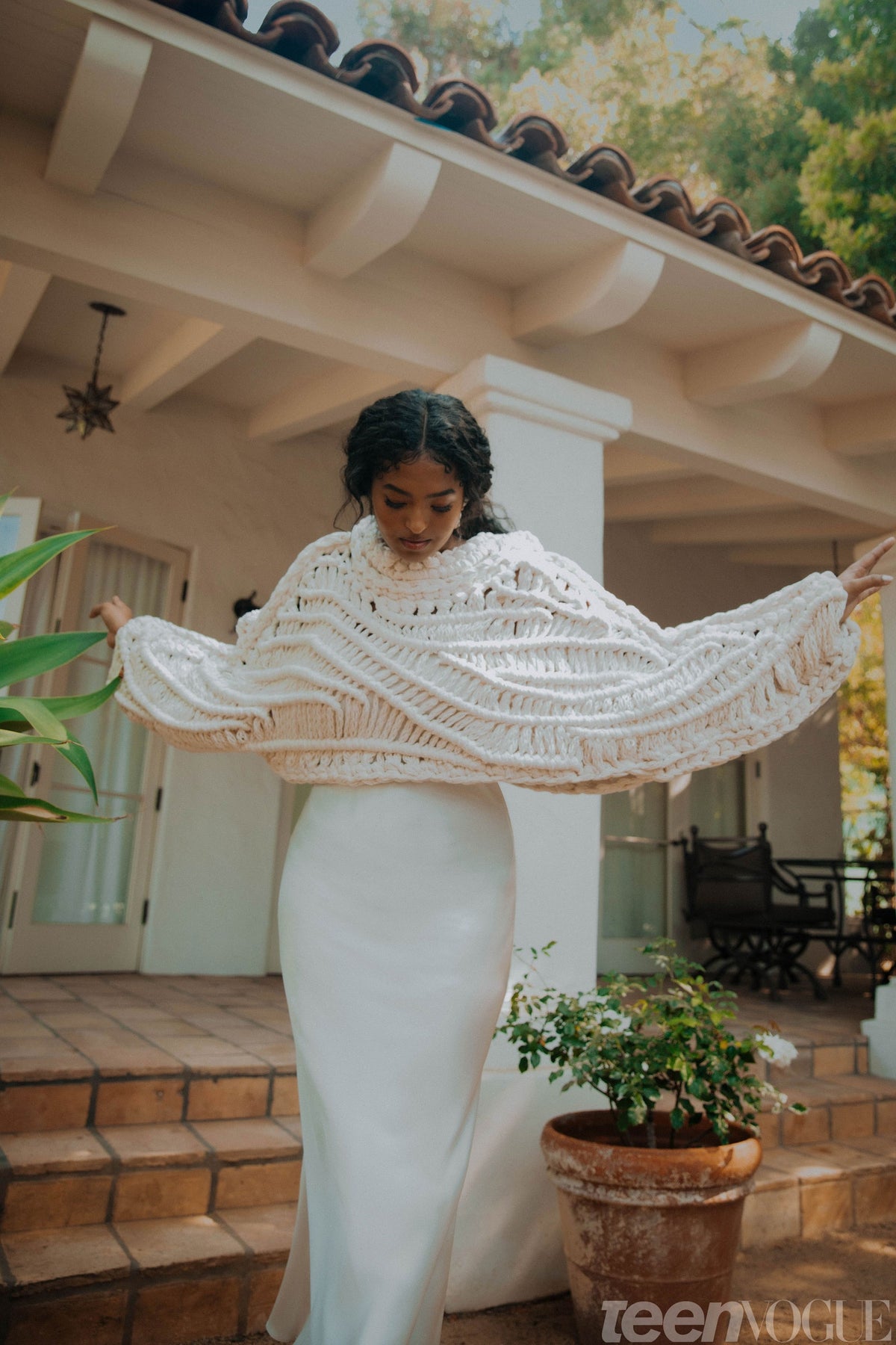 A model wearing a knitted shawl over a white dress, standing in a garden.