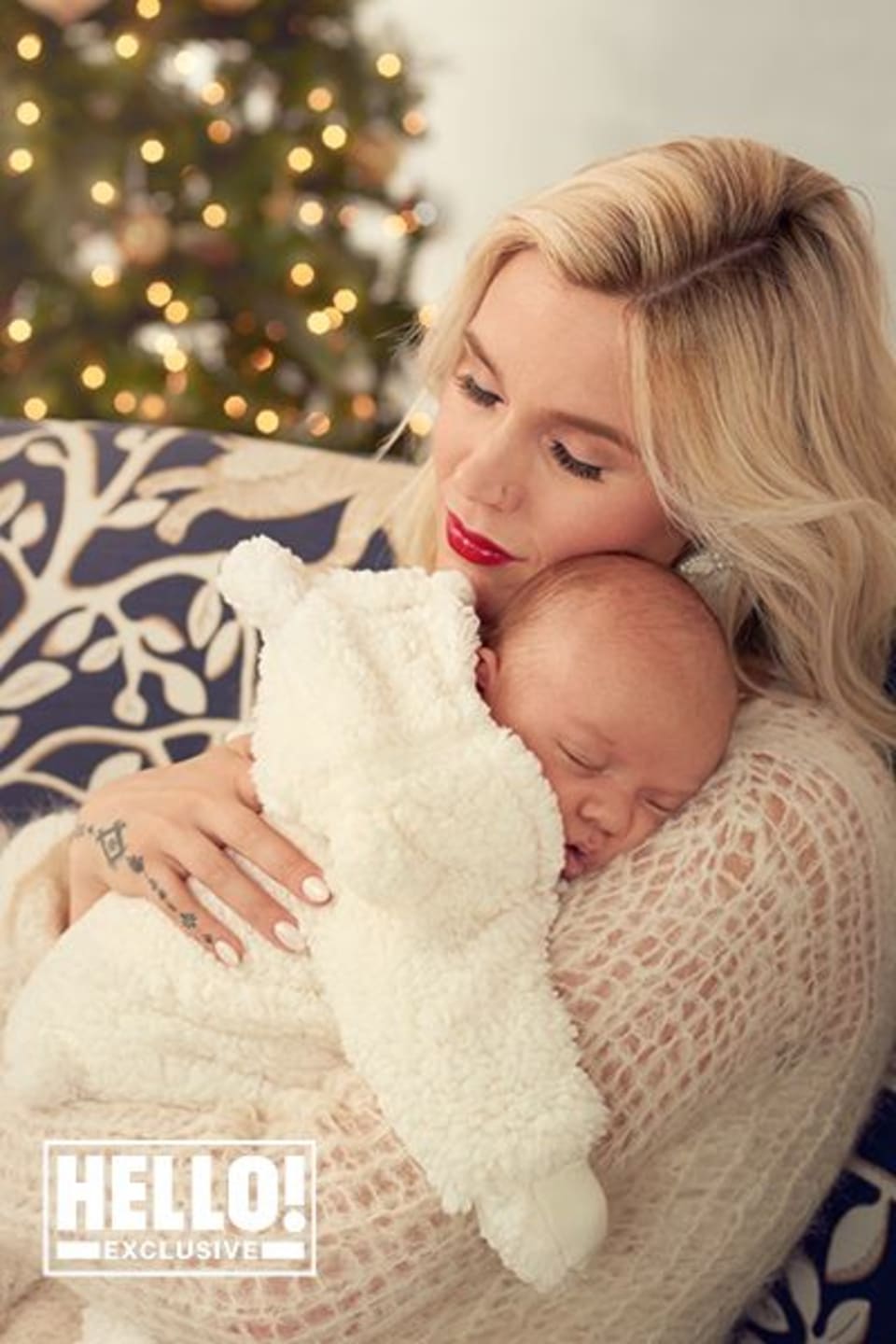 A woman with blonde hair holding a sleeping baby in a cozy setting with holiday decorations in the background.